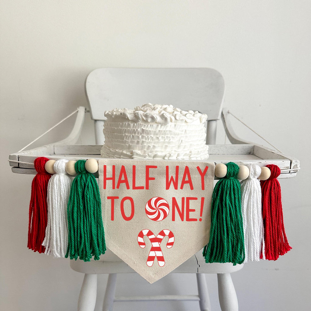 A 6 by 7 inch canvas center flag with the words HALFWAY TO ONE in red with a peppermint as the O. There are 2 criss crossed candy canes under the words. There is a 5 inch green, white and red yarn tassel on each side each separated by a wooden bead.