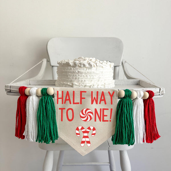 A 6 by 7 inch canvas center flag with the words HALFWAY TO ONE in red with a peppermint as the O. There are 2 criss crossed candy canes under the words. There is a 5 inch green, white and red yarn tassel on each side each separated by a wooden bead.