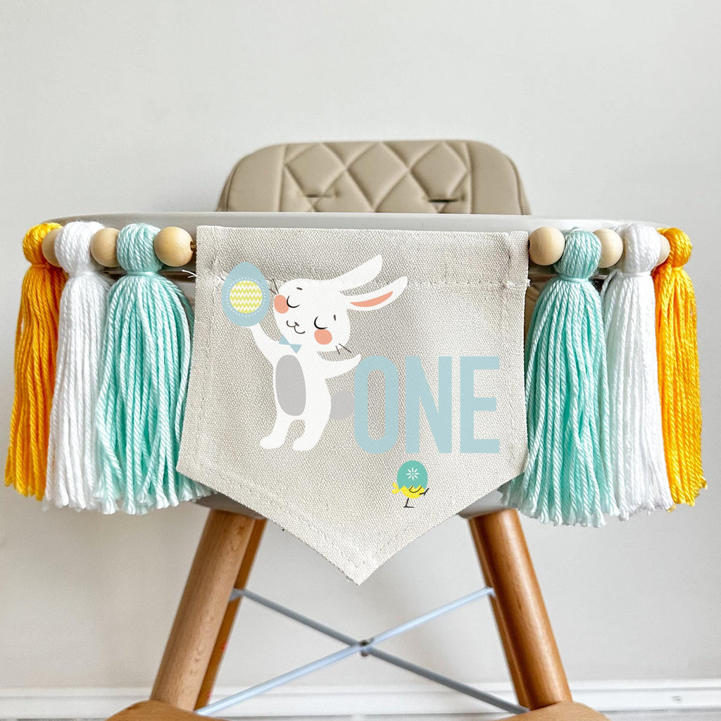 A 5 by 7 inch canvas center flag with a mint number 1 and a bunny peeking around it with a garden of Easter eggs at the bottom. There is a 5 inch mint, white and yellow tassel on either side, each separated by a wooden bead for a total of 6 tassels.
