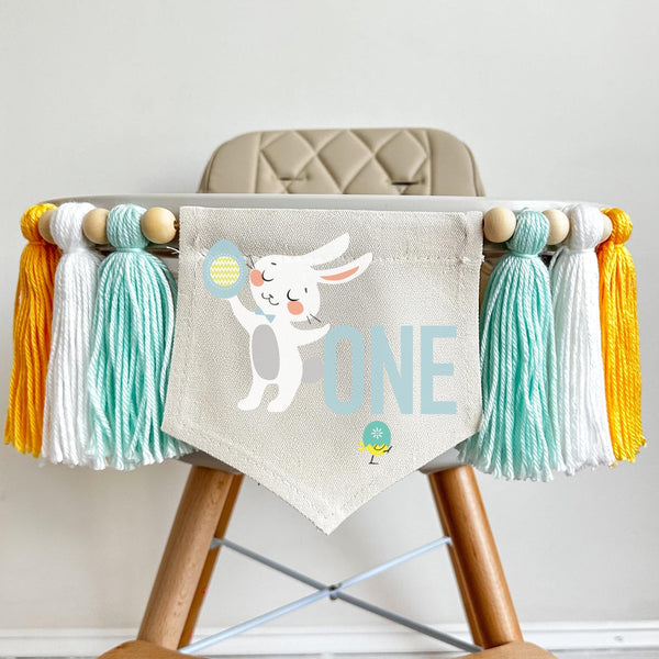 A 5 by 7 inch canvas center flag with a mint number 1 and a bunny peeking around it with a garden of Easter eggs at the bottom. There is a 5 inch mint, white and yellow tassel on either side, each separated by a wooden bead for a total of 6 tassels.
