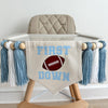 A 5 by 7 inch canvas center flag with the words FIRST DOWN in light blue with a football in the middle. There is a 5 inch light blue, white and light blue yarn tassel on each side each separated by a wooden bead for a total of 6 tassels.