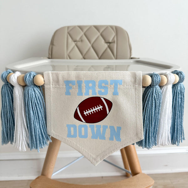 A 5 by 7 inch canvas center flag with the words FIRST DOWN in light blue with a football in the middle. There is a 5 inch light blue, white and light blue yarn tassel on each side each separated by a wooden bead for a total of 6 tassels.