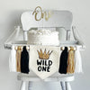 A 5 by 7 inch canvas banner with the words WILD ONE in black and a gold sparkly printed crown above the words. There is a 5 inch black, tan and white yarn tassel on each side with a wooden bead between each of them for a total of 6 tassels.