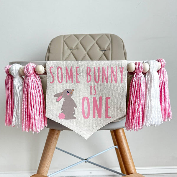 A 5 by 7 inch canvas center flag with the words SOME BUNNY IS ONE in pink with a grayish brown bunny with pink features beside the words. On each side there is a pink, white and pink 5 in yarn tassel each separated by a wooden bead for a total of 6.