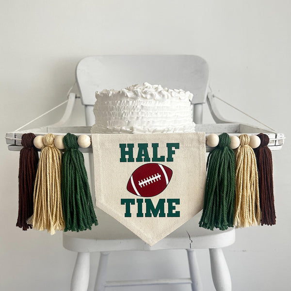 A 5 by7 inch canvas flag with the words HALF TIME in dark green with a football in the middle. There is a 5 inch dark green, tan and brown yarn tassel on each side, each separated by a wooden bead for a total of 6 tassels. It is hung on a high chair.
