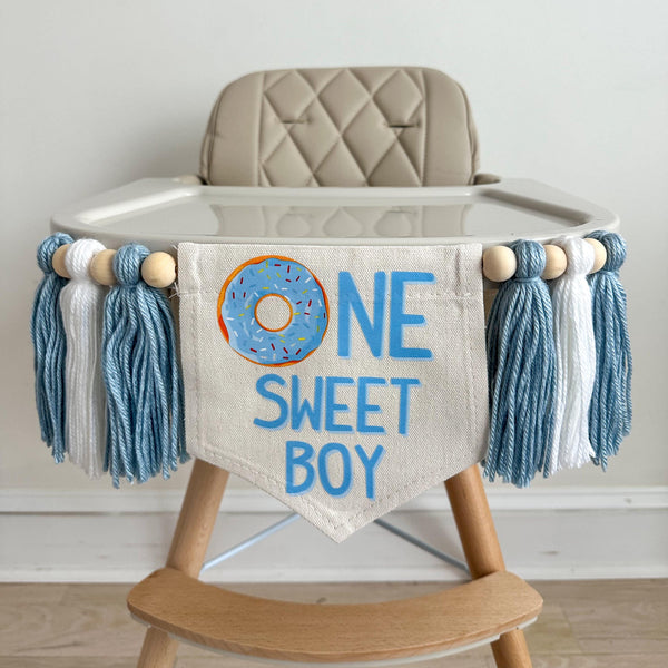 A 5 by 7 inch canvas center flag with the words ONE SWEET BOY in light blue with a blue donut as the O. There are 5 inch yarn tassels on each side in light blue, white and light blue each separated by a wooden bead. There are a total of 6 tassels.