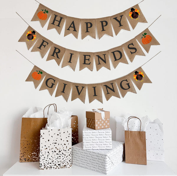 A burlap banner made from 4.5 by 6.5 inch fishtail shaped flags on 3 strings. The top line says HAPPY in black capital letters with a hand stenciled pumpkin and a hand stenciled turkey on each end. The middle string says FRIENDS with a turkey and a pumpkin on each end. The bottom string says GIVING with a pumpkin and a turkey on each end.