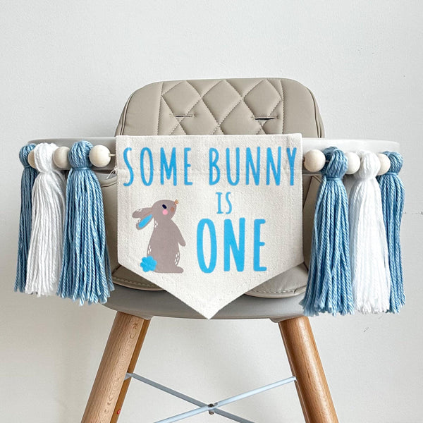 A 5 by 7 inch canvas center flag with the words SOME BUNNY IS ONE in baby blue with a grayish brown bunny with blue features beside the words. On each side there is a light blue, white and light blue 5 in yarn tassel each separated by a wooden bead.