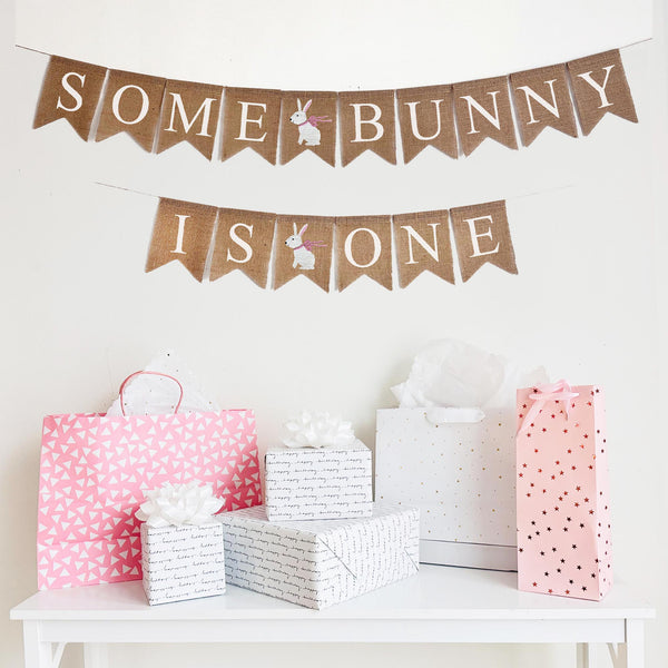 A burlap banner split into two lines. The first line reads &quot;Some Bunny&quot; with a cute hand stenciled white bunny in a pink bow in between the words. The second line reads &quot;Is One&quot; with a hand stenciled white bunny with a pink bow in between the words. The perfect addition to a spring first birthday celebration.