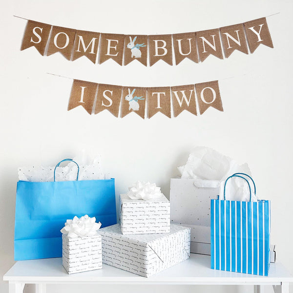 Some Bunny is Two burlap banner in blue. This banner is in two rows, the top row says &quot;Some Bunny&quot; with a white hand stenciled bunny wearing a blue ribbon in between the words. The second line reads &quot;Is Two&quot; with a hand stenciled bunny flag in between the words.