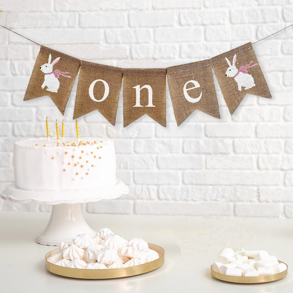 A bunny themed one banner on burlap fishtail flags. This banner starts with one white hand stenciled bunny wearing a pink bow followed by the words one in white lower case letters and ends with another white hand stenciled bunny image wearing a pink bow.