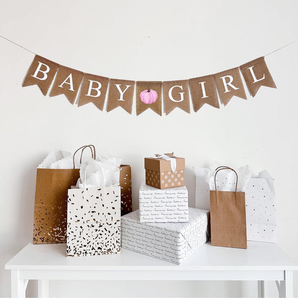 A burlap banner made from 4.5 by 6.5 inch fishtail shaped flags. Each flag has a pocket across the top for stringing. This banner says BABY GIRL in white capital letters. There is a printed image of a pink watercolor pumpkin with a brown stem between each word. It is strung on a jute string that is 110 inches in length.