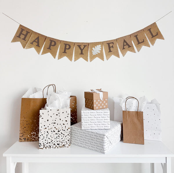 A burlap banner made from 4.5 by 6.5 inch fishtail shaped flags. Each flag has a pocket across the top for stringing. This banner says HAPPY FALL in brown capital letters. There is a hand stenciled image of a white oak leaf in between each word. It is strung on a jute string that is 110 inches in length.