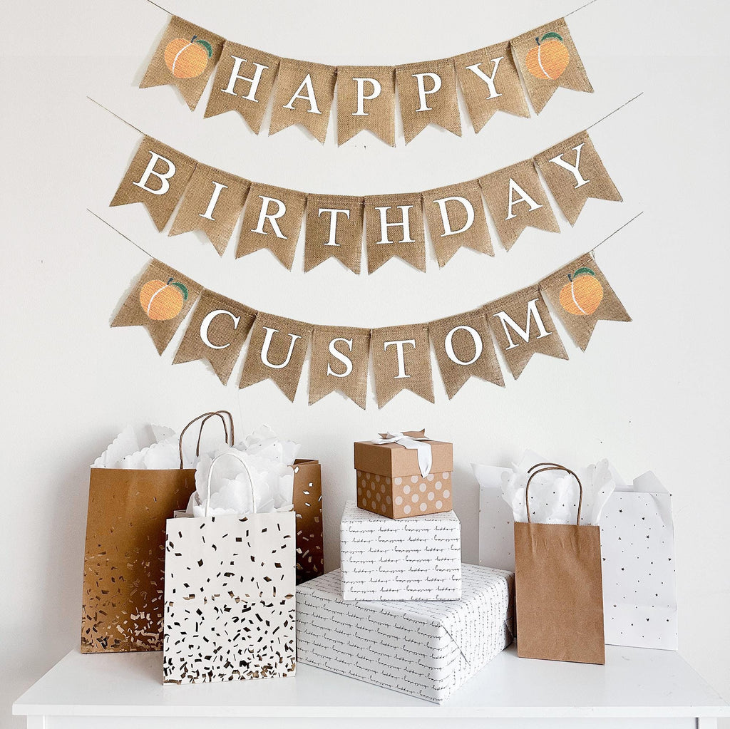 This is a burlap banner made out of 4 inch by 6 inch fishtail flags. It is on three strings. The top string has the word Happy in white letters with an image of a peach on either end. The second string has the word Birthday in white letters. The top string has the word CUSTOM in white letters with an image of a peach on either end.