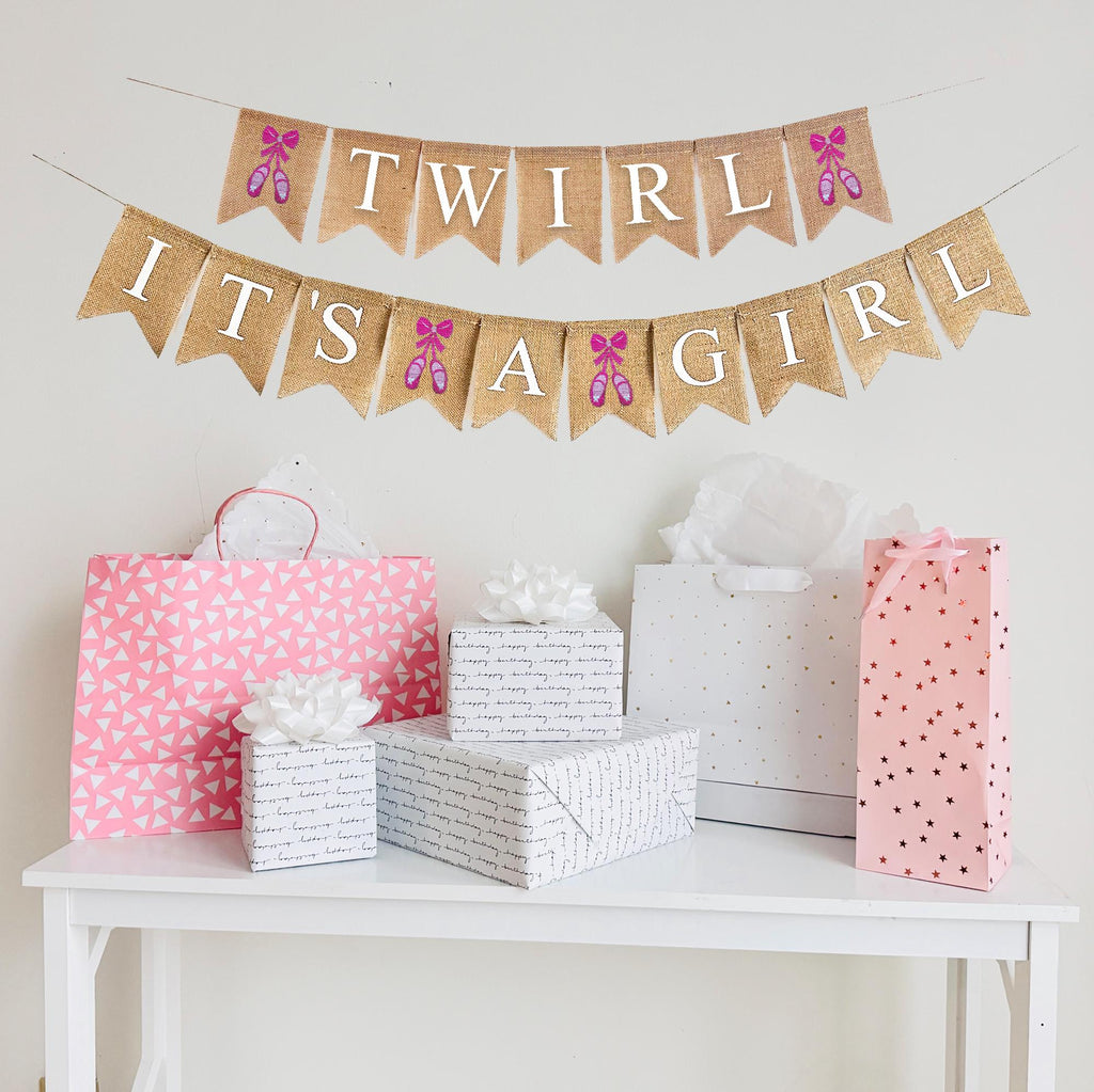 A burlap banner made from 4.5 by 6.5 inch fishtail shaped flags. Each flag has a pocket across the top for stringing. This banner says TWIRL on the top string in white capital letters. It has a hand stenciled image of pink ballet shoes on each end of the word. The bottom string says IT&#39;S A GIRL in white capital letters. There is a hand stenciled image of a pair of pink ballet shoes between each word. Each line is strung on a jute string that is 110 inches in length.