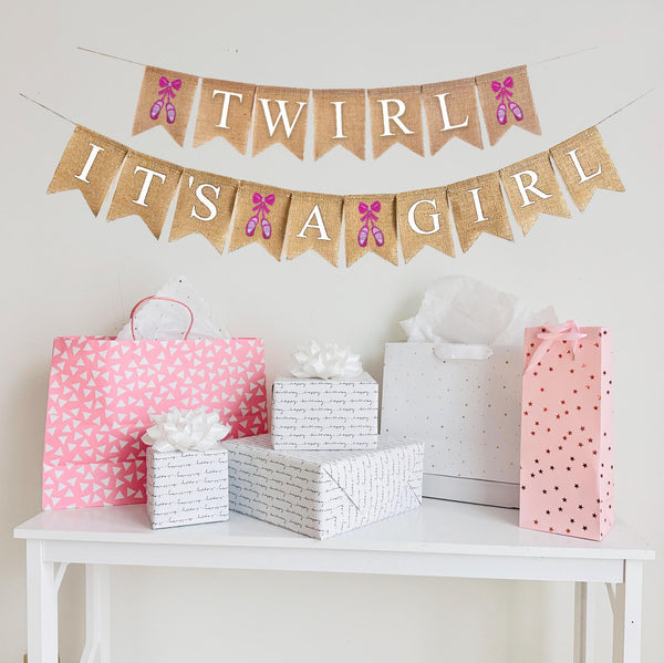 A burlap banner made from 4.5 by 6.5 inch fishtail shaped flags. Each flag has a pocket across the top for stringing. This banner says TWIRL on the top string in white capital letters. It has a hand stenciled image of pink ballet shoes on each end of the word. The bottom string says IT&#39;S A GIRL in white capital letters. There is a hand stenciled image of a pair of pink ballet shoes between each word. Each line is strung on a jute string that is 110 inches in length.