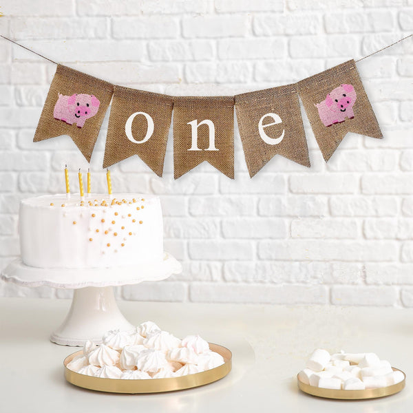 This farm themed first birthday burlap banner is make up of 5 burlap flags. The first flag is a hand stenciled baby pig followed by three more flags spelling out the word one in lowercase white letters. The banner ends with another hand stenciled baby pig.