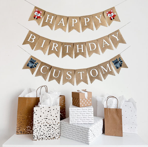 A burlap banner made from 4.5 by 6.5 inch fishtail shaped flags. Each flag has a pocket across the top for stringing. This is a custom banner in white capital letters. The top string says HAPPY and has a hand stenciled image of a fox on each end. The second string says BIRTHDAY with no images. The bottom line can be customized with a name and has a hand stenciled image of a raccoon on each end. It is strung on a jute string that is 110 inches in length.