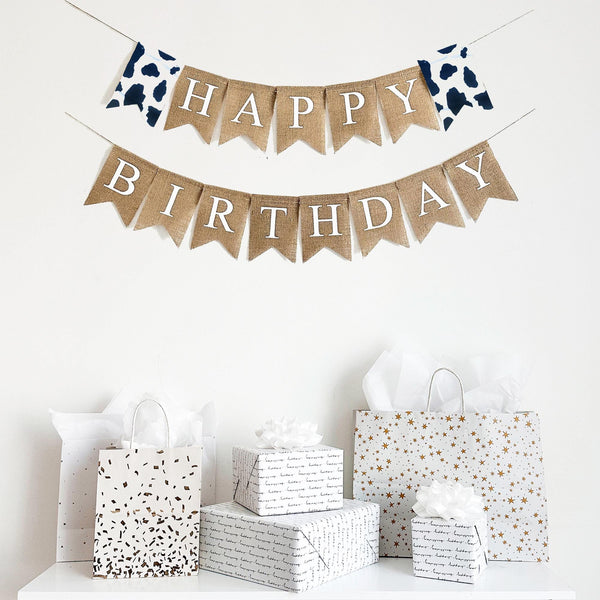 Happy Birthday banner made of cow print fabric and burlap. The first line of this banner starts with a cow print flag and reads Happy followed by another cow print flag. The second line reads Birthday. All the words are in white letters on top of a beige burlap flag. This banner is customizable and the perfect addition to a farm party theme.