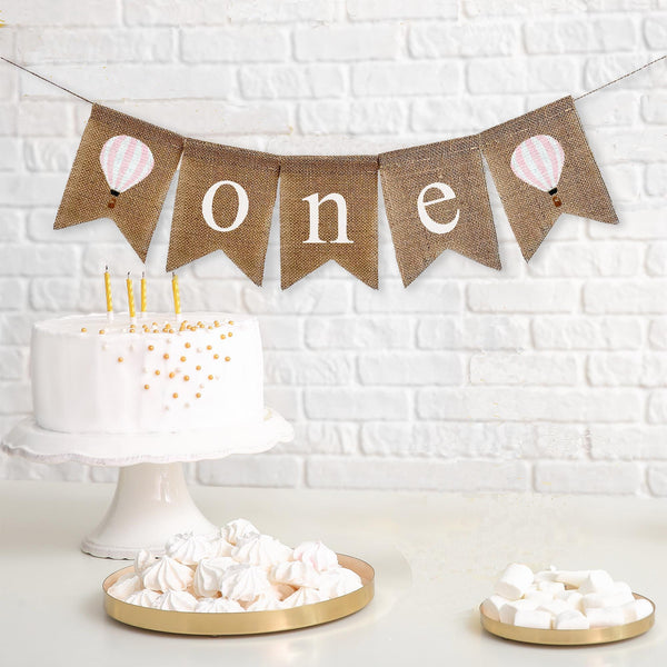 A burlap banner with letters reading &quot;one&quot; with images of white and pink striped hot air balloons on each side of the word.