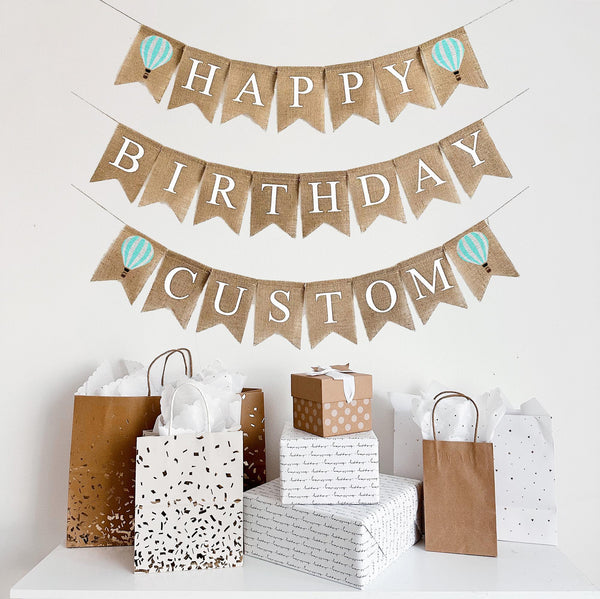 A happy birthday burlap banner strung on three strings. The happy is at the top and the birthday below with CUSTOM on the bottom.  Hand painted mint and white striped hot air balloons are at the beginning and end of the word happy and custom.
