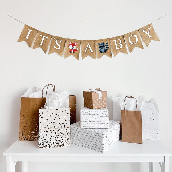 A burlap banner made from 4.5 by 6.5 inch fishtail shaped flags. Each flag has a pocket across the top for stringing. The banner says IT&#39;S A BOY in white capital letters. There is a hand stenciled image of a fox between the first two words and a hand stenciled image of a raccoon between the second two words.  It is strung on a jute string that is 110 inches in length.