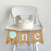 A burlap banner with a mint and white striped hot air balloon and the letters n and e, making the banner spell one with the hot air balloon as the o.