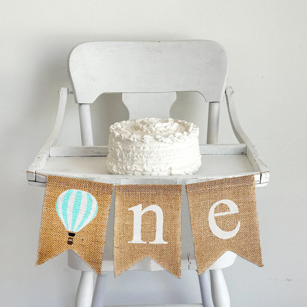 A burlap banner with a mint and white striped hot air balloon and the letters n and e, making the banner spell one with the hot air balloon as the o.