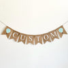 A burlap banner with the wording CUSTOM between two mint & white striped hot air balloon image flags.