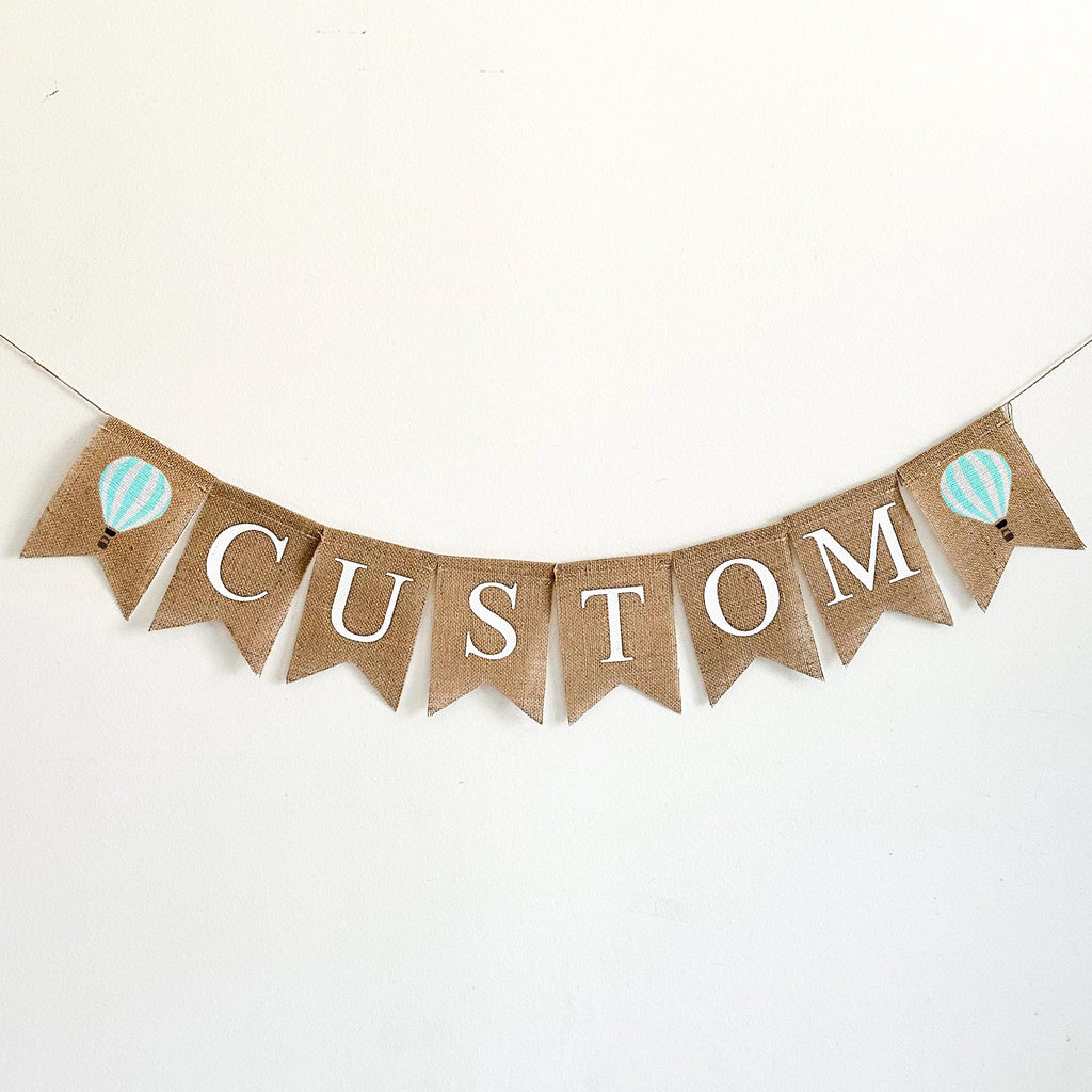 A burlap banner with the wording CUSTOM between two mint & white striped hot air balloon image flags.