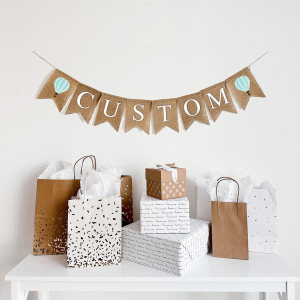 A burlap banner with the wording CUSTOM between two mint & white striped hot air balloon image flags.