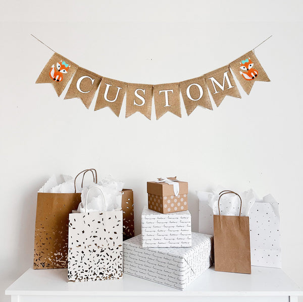 A burlap banner made from 4.5 by 6.5 inch fishtail shaped flags. Each flag has a pocket across the top for stringing. This is a custom banner in white capital letters. Add the name of your choosing to personalize this banner. There is a hand stenciled image of an orange girl fox with a bow on each end of the name. It is strung on a jute string that is 110 inches in length.
