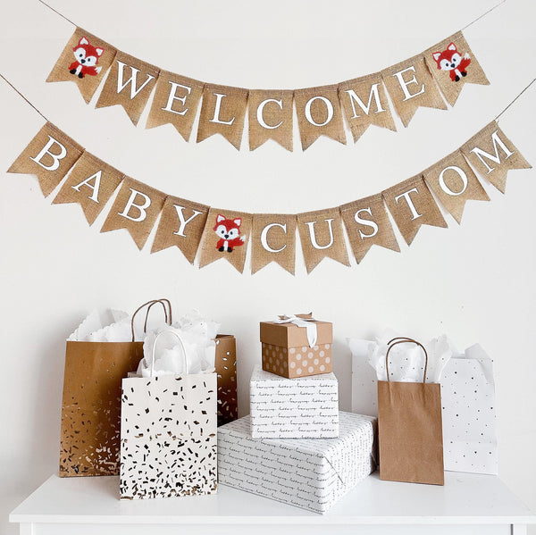 A burlap banner made from 4.5 by 6.5 inch fishtail shaped flags. Each flag has a pocket across the top for stringing. The top line says WELCOME in white capital letters. There is a hand stenciled image of a fox on each end. The bottom string says BABY CUSTOM and has a hand stenciled image of a fox between the words. Add your own name to this banner. It is strung on two jute strings that are 110 inches in length.
