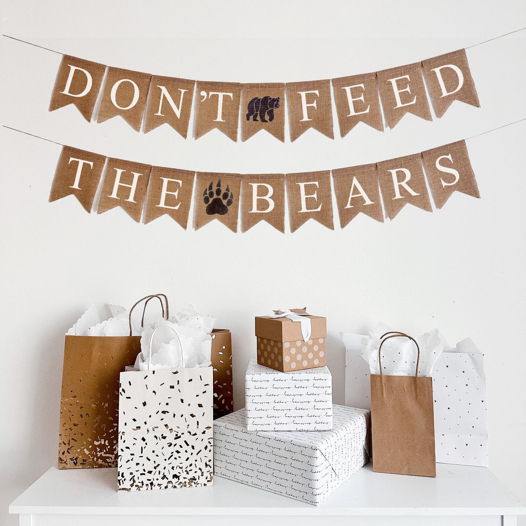 A burlap banner made from 4.5 by 6.5 inch fishtail shaped flags. Each flag has a pocket across the top for stringing. The top string of this banner says DON&#39;T FEED in white capital letters with a hand stenciled image of a bear in between the words. The bottom string says THE BEARS in white capital letters with a hand stenciled brown bear paw print in between the words. It is strung on two jute strings that are 110 inches in length.