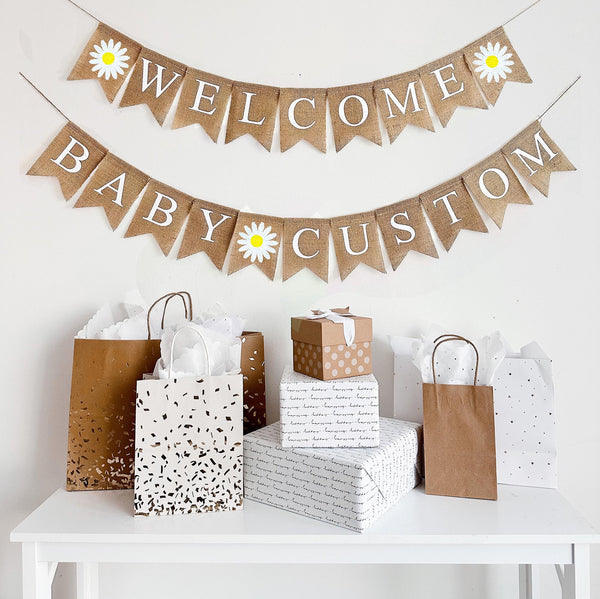 A burlap banner made from 4.5 by 6.5 inch fishtail shaped flags. Each flag has a pocket across the top for stringing. The top line says WELCOME in white capital letters. There is a hand stenciled image of a daisy with a yellow center on each end. The bottom string says BABY CUSTOM and has a hand stenciled image of a daisy between the words. Add your own name to this banner. It is strung on two jute strings that are 110 inches in length.
