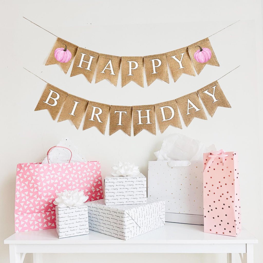 A burlap banner made from 4.5 by 6.5 inch fishtail shaped flags. The top string says HAPPY in white capital letters with a pink printed watercolor pumpkin on each end. The bottom string says BIRTHDAY in white capital letters.