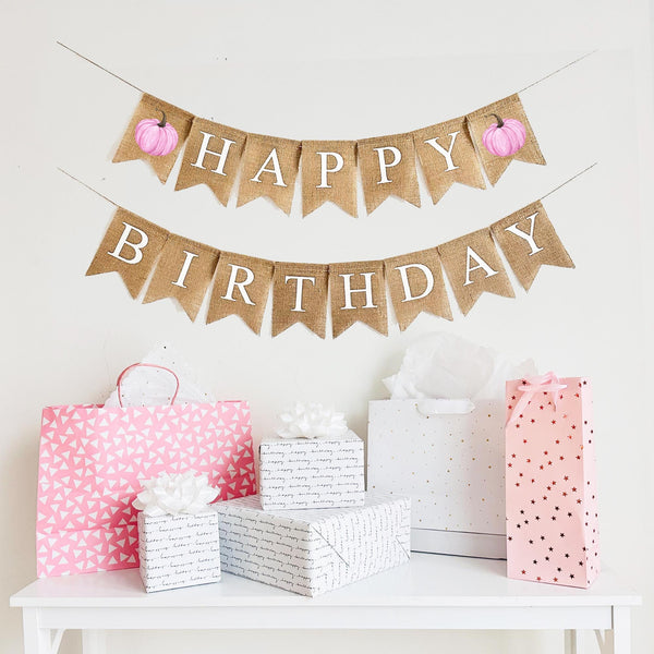 A burlap banner made from 4.5 by 6.5 inch fishtail shaped flags. The top string says HAPPY in white capital letters with a pink printed watercolor pumpkin on each end. The bottom string says BIRTHDAY in white capital letters.