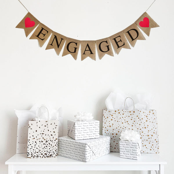 Burlap banner that spells out the word Engaged in black letters on a burlap fishtail banner. Each end of the banner had a bright red heart. Celebrate love with this classic burlap banner.