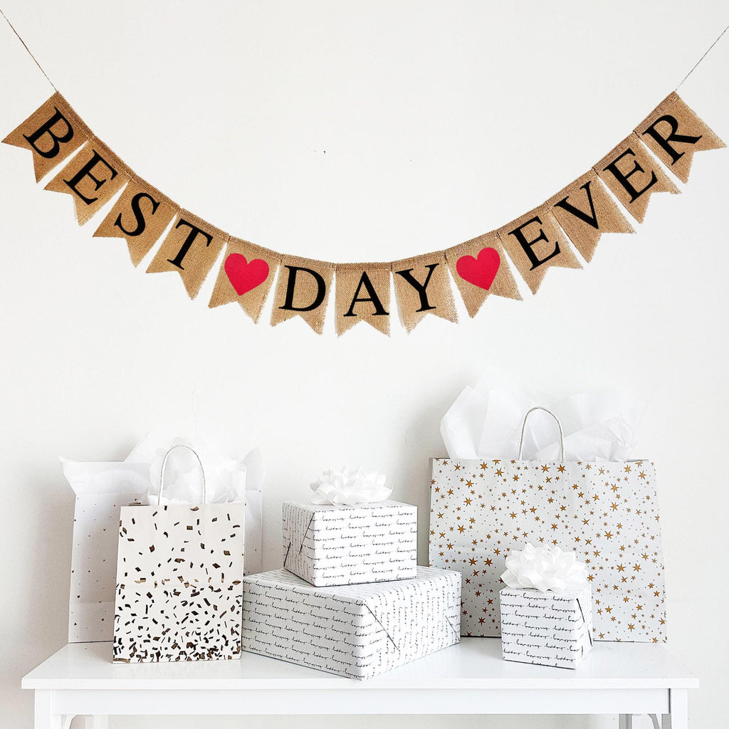 This banner consists of fishtail burlap flags with black letters that spell out the words BEST DAY EVER. In between each word is a red heart on a burlap fishtail flag. There are 2 red hearts in total. This banner is perfect for a wedding photo booth back drop!