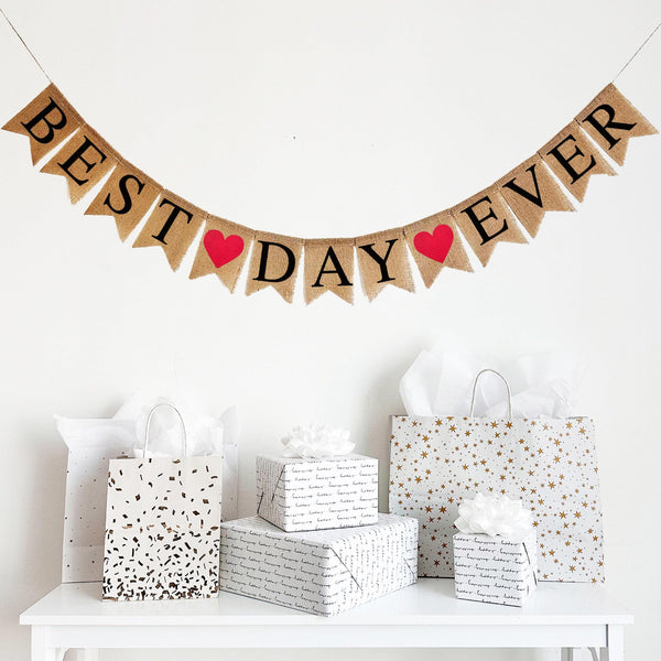 This banner consists of fishtail burlap flags with black letters that spell out the words BEST DAY EVER. In between each word is a red heart on a burlap fishtail flag. There are 2 red hearts in total. This banner is perfect for a wedding photo booth back drop!