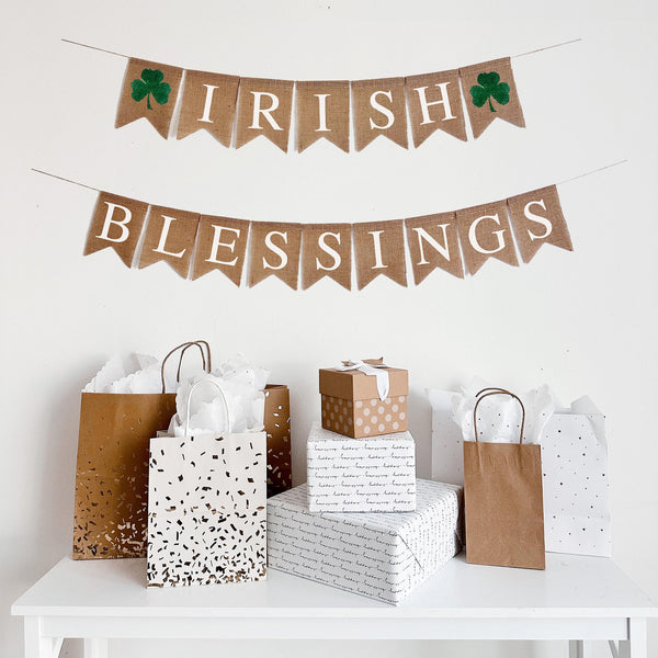Irish Blessings burlap banner featuring 2 hand stenciled green sharmrocks. This banner is 2 rows, on the first row there is a shamrock flag followed by flags spelling out Irish and another shamrock at the end. The second line, the word blessings is spelled out in white font, one letter stenciled on each burlap flag. This banner is the perfect addition to your St. Patrick&#39;s Day decor.