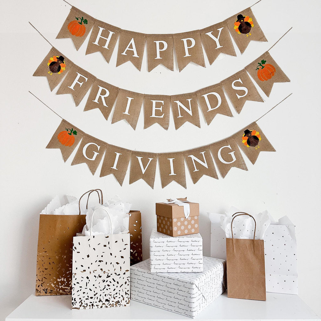A burlap banner made from 4.5 by 6.5 inch fishtail shaped flags on 3 strings. The top line says HAPPY in white capital letters with a hand stenciled pumpkin and a hand stenciled turkey on each end. The middle string says FRIENDS with a turkey and a pumpkin on each end. The bottom string says GIVING with a pumpkin and a turkey on each end.