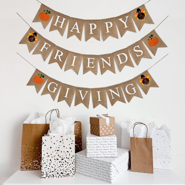 A burlap banner made from 4.5 by 6.5 inch fishtail shaped flags on 3 strings. The top line says HAPPY in white capital letters with a hand stenciled pumpkin and a hand stenciled turkey on each end. The middle string says FRIENDS with a turkey and a pumpkin on each end. The bottom string says GIVING with a pumpkin and a turkey on each end.