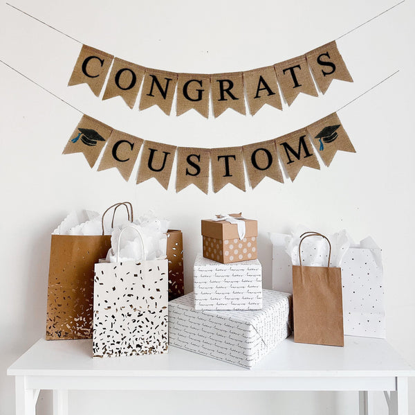 A burlap banner made from 4.5 by 6.5 inch fishtail shaped flags. Each flag has a pocket across the top for stringing. This is a custom banner in black capital letters. The top line says CONGRATS in black capital letters. Add the name or word of your choosing to the second line to personalize this banner. There is a hand stenciled image of a black graduation cap with a blue tassel on each end of the name. It is strung on a jute string that is 110 inches in length.