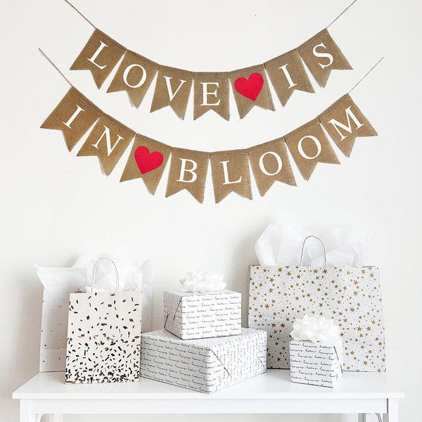 A two line burlap banner with white letters spelling out the words Love Is on the first line and In Bloom on the second line. There is a bright red heart between the words Love and Is and another red heart between in and bloom making this a perfect love filled banner design.