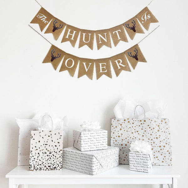 This burlap fishtail flag banner is hung in two lines. The top line starts with a flag with the words The stenciled in script lettering followed by a flag with a hand stenciled brown deer head silhouette. The end of the banner has another hand stenciled deer head followed by the word Is stenciled in script font on one flag. The full first line reads &quot;The Hunt Is&quot; The second line begins with another deer image followed by the word &quot;Over&quot; and then another deer stencil.