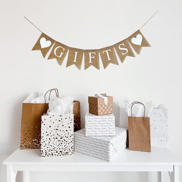 A burlap banner made from 4.5 by 6.5 inch fishtail shaped flags. Each flag has a pocket across the top for stringing. This banner says GIFTS in white capital letters. There is a hand stenciled image of a white heart on each end of the word. It is strung on a jute string that is 110 inches in length.