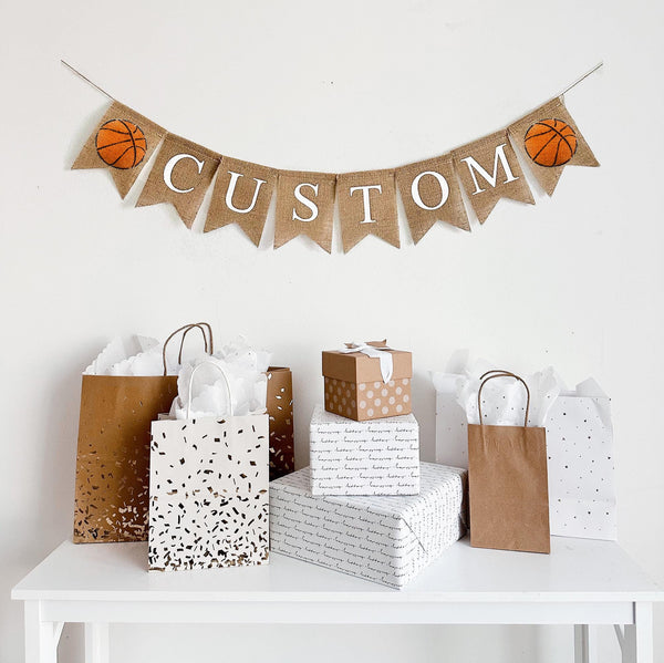 A burlap banner made from 4.5 by 6.5 inch fishtail shaped flags. Each flag has a pocket across the top for stringing. This is a custom banner in white capital letters. Add the name of your choosing to personalize this banner. There is a hand stenciled image of an orange basketball on each end of the name. It is strung on a jute string that is 110 inches in length.