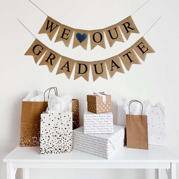 A burlap banner made from 4.5 by 6.5 inch fishtail shaped flags. Each flag has a pocket across the top for stringing. The top line says WE heart OUR in black capital letters with a navy heart in between the words instead of the word love. The bottom line says GRADUATE in black capital letters. Each line is strung on a jute string that is 110 inches in length.