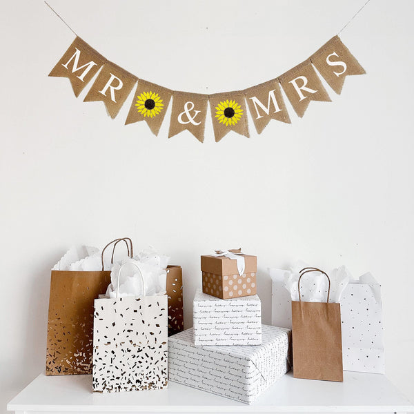 A burlap banner reading Mr & Mrs with a hand stenciled sunflower image flag between the words. Each burlap flag measures 4.5 x 6.5 and the words are written in all capital letters in white lettering. The sunflower has a dark brown center and yellow petals.